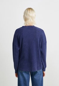 Pull over long à manches longues oversize bleu marine en tissu doux et texturé. Présente des épaules tombantes et un col en rib, associé à un jean en denim.