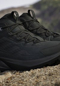 Czarne buty trekkingowe mają teksturowaną cholewkę, wzmocniony czubek oraz grubą gumową podeszwę z wyraźnym wzorem bieżnika. Sznurowadła i oczka są w zestawie.
