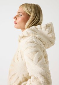 Giacca con cappuccio di colore crema realizzata in pile testurizzato, con un motivo a foglia prominente sulla superficie del tessuto e un design con cappuccio.