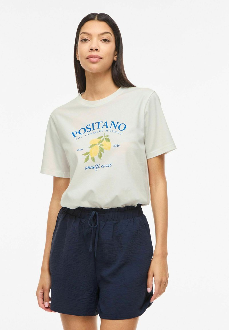 Femme portant un t-shirt blanc avec un motif citron et le texte "Positano Amalfi Coast", associé à un short bleu marine à cordon de serrage.