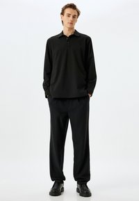 Schwarzes Langarm-Polohemd mit Kragen und drei Knöpfen, kombiniert mit locker sitzenden schwarzen Hosen und klobigen schwarzen Schuhen.