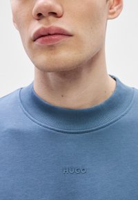 Μπλε φούτερ με ριμπ λαιμόκοψη, με ανάγλυφο λογότυπο "HUGO" στο στήθος. Λείο ύφασμα και κοντινή λήψη της λαιμόκοψης και της περιοχής του προσώπου.
