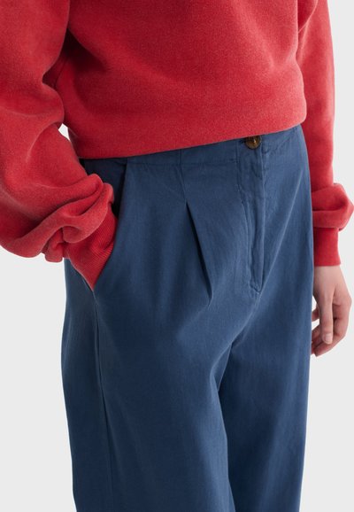 Personne portant un haut rouge à manches longues et un pantalon taille haute bleu, les mains partiellement dans les poches, sur un fond uni.
