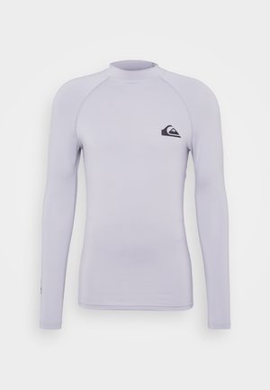 Rash guard à manches longues en tissu lavande clair, présentant un design ajusté, un col rond et un petit logo noir sur la poitrine.