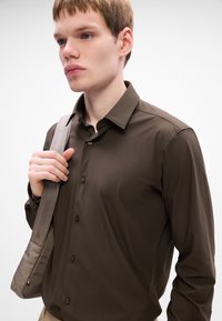 Camisa de botones de manga larga color marrón oscuro con cuello, con botones en la parte delantera y tejido suave, combinada con un bolso de hombro gris claro.