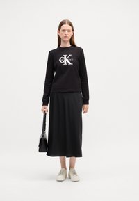 Juodas ilgas rankovėmis marškinėlis su baltu "CK" logotipu, derinamas su ilga juoda sijonu ir baltais sportbačiais, laiko juodą rankinę. Skaidrios medžiagos.