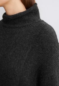 Pull en tricot gris foncé avec un col haut plié, présentant un motif texturé et une coupe ample, mettant l'accent sur la chaleur et le confort.