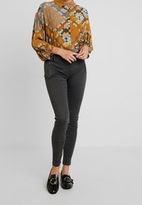 Långärmad skjorta med krage i guld och flerfärgade geometriska mönster, tillsammans med högmidjade, mörkgrå skinny jeans och svarta loafers med ringdetalj.