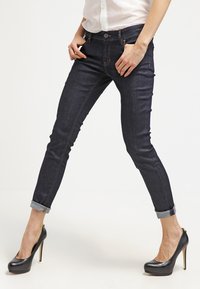 Jeans skinny de mezclilla azul oscuro con puños enrollados, que cuentan con un cierre delantero de botón y costuras en contraste. Combinados con tacones negros.