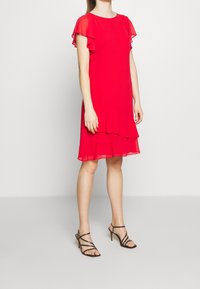 Femme portant une robe rouge jusqu'aux genoux avec des manches courtes ondulées et un ourlet, associée à des sandales à talons hauts noires avec des lanières, sur un fond uni.