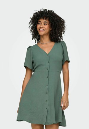 Femme aux cheveux bouclés souriante, portant une robe verte à manches courtes avec des boutons sur le devant et un collier en argent, debout devant un fond uni.