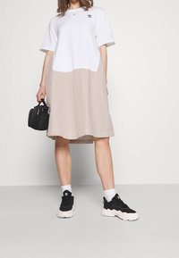 T-paita mekko, jossa on valkoinen yläosa ja raitainen beige hame. Lyhyet hihat, rento malli ja mustat tennarit. Pieni laukku kädessä.