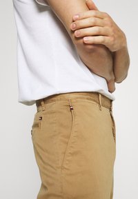 Pantalons slim-fit beige en coton avec une texture lisse, dotés d'une fermeture éclair, de poches latérales et d'accents logo décoratifs sur la ceinture.