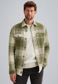 Groen en crèmekleurig geruite jas met knoopsluiting, twee borstzakken en textielstructuur; gedragen over een crèmekleurige trui en zwarte spijkerbroek.