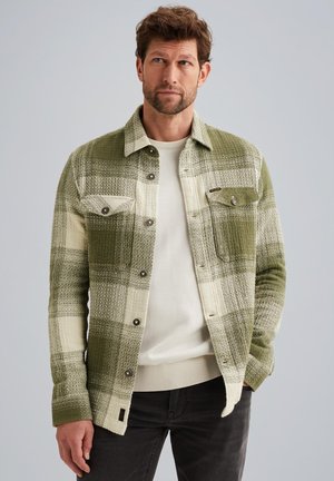 Groen en crèmekleurig geruite jas met knoopsluiting, twee borstzakken en textielstructuur; gedragen over een crèmekleurige trui en zwarte spijkerbroek.