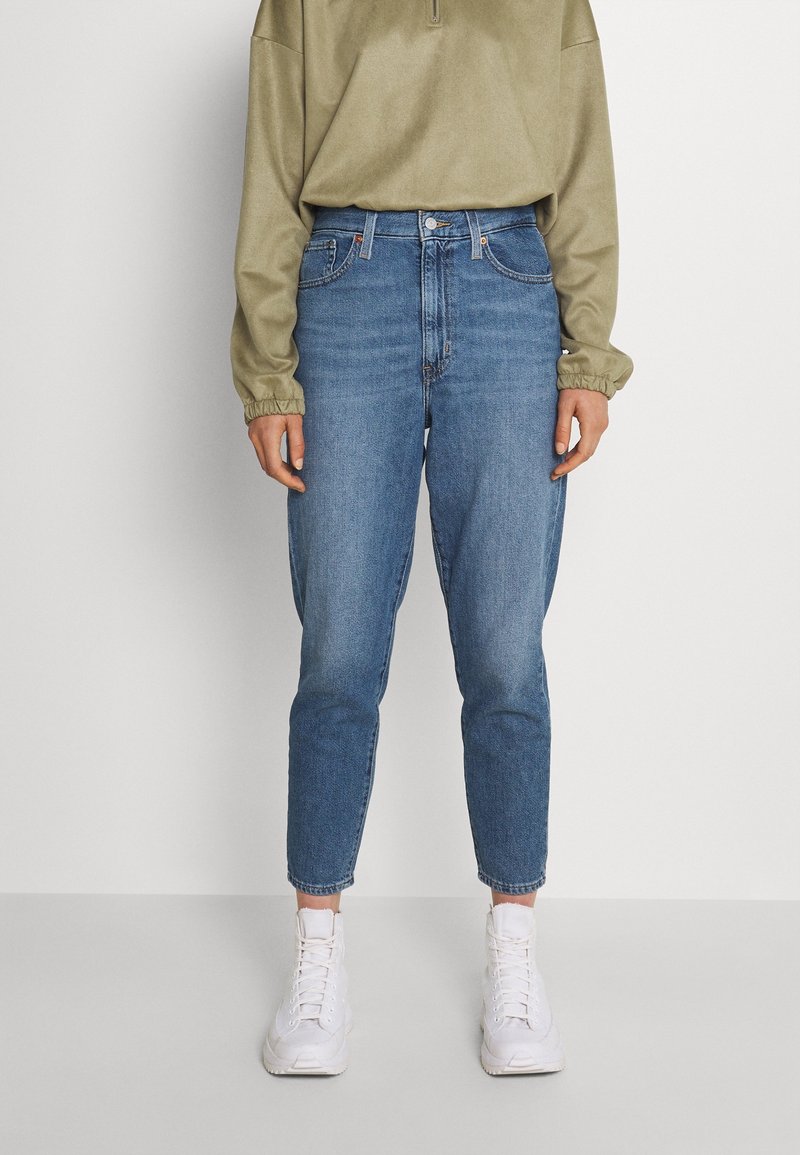 Jean denim bleu taille haute avec jambes fuselées, présentant une décoloration subtile, un design à cinq poches et une fermeture à boutons, assorti à un haut vert.