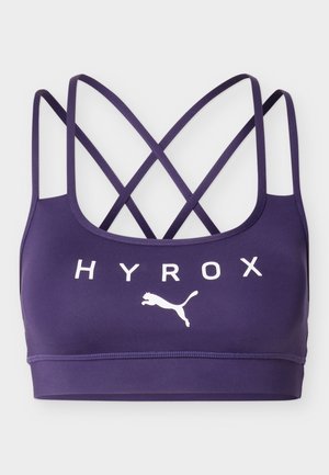 Sujetador deportivo morado con tirantes cruzados y texto blanco HYROX junto con el logo de Puma en el frente sobre un fondo claro.