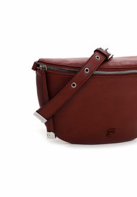 Rød læder crossbody taske med lynlås, justerbar strop, sølvhardware og prægede logo detaljer. Glat tekstur, rund form.