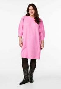 Abito rosa lavorato a maglia con maniche a sbuffo, polsini e orlo a costine. Indossato sopra collant neri e stivali cowboy neri con punta caratterizzati da dettagli con borchie.