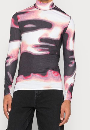 Uomo che indossa una maglia a maniche lunghe con collo alto e stampa astratta di un volto in toni di nero, bianco e rosa, abbinata a pantaloni neri.