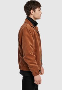 Giacca in velluto a coste marrone con chiusura frontale a cerniera, bordo e polsini a coste e colletto, indossata da un modello di profilo su uno sfondo neutro.