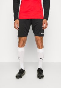 Schwarze Sportshorts mit einem weißen "JAKO"-Logo, kombiniert mit weißen Socken, die ein schwarzes Puma-Logo tragen, und schwarzen Fußballschuhen.