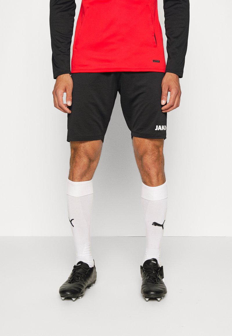 Schwarze Sportshorts mit einem weißen "JAKO"-Logo, kombiniert mit weißen Socken, die ein schwarzes Puma-Logo tragen, und schwarzen Fußballschuhen.