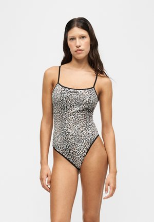 Woman with long dark hair wearing a leopard print one-piece swimsuit with thin black straps, standing against a plain light background.