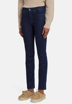 Vrouw draagt donkerblauwe skinny jeans, beige trui en beige casual veterschoenen, staat met handen langs het lichaam.