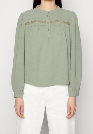 Kvinde iført lysegrøn bluse med lange ærmer, knaplukning ved halsen og blonde detaljer, kombineret med hvide bukser med højtaljet pasform.