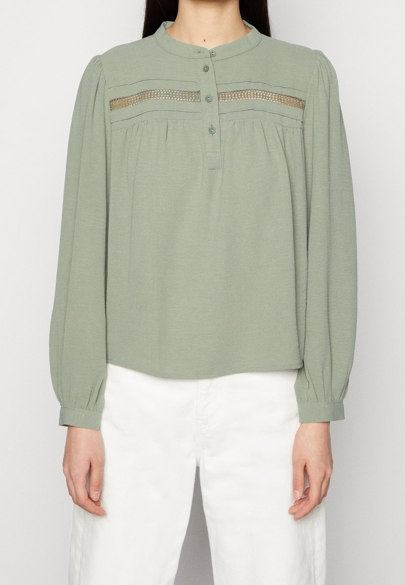 Femme portant un chemisier vert clair à manches longues avec un col boutonné et un détail en dentelle, associé à un pantalon blanc taille haute.