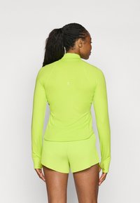 Limettengrünes Sportoberteil mit hohem Kragen und langen Ärmeln, kombiniert mit passenden gerippten Shorts. Glatte Textur, minimalistisches Design.