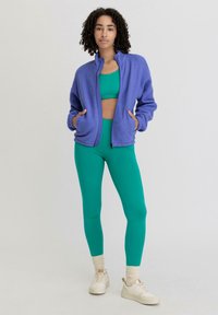Fleecejacke in Violett mit hohem Kragen, kombiniert mit einem grünen Crop-Top und Leggings. Weiße Sneakers vervollständigen das Outfit.