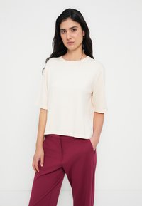 Ανοιχτό μπεζ τοπ με κοντά μανίκια και στρογγυλή λαιμόκοψη, κατασκευασμένο από μαλακό ύφασμα. Συνδυάζεται με ψηλόμεσα βυσσινί παντελόνια, με πλαϊνές τσέπες.