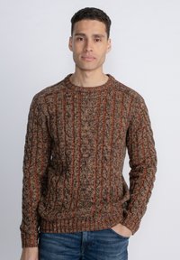 Brown and orange knitted sweater with a textured cable pattern, crew neckline, and ribbed cuffs and hem, worn with blue jeans.