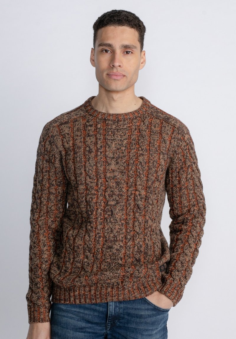 Brown and orange knitted sweater with a textured cable pattern, crew neckline, and ribbed cuffs and hem, worn with blue jeans.