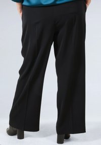 Person wearing wide-leg black trousers and black heeled shoes, standing with hands in pockets against a plain background.