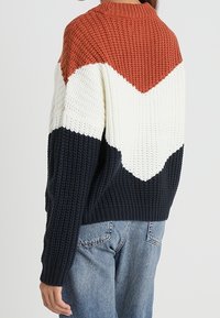 Sweter dziany z wzorem w kolorowe bloki w odcieniach rdzy, bieli i granatu, o fakturze prążkowanej i klasycznym okrągłym dekolcie.