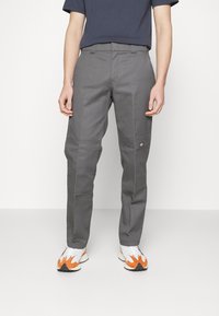 Homme portant un pantalon droit gris avec des genoux renforcés et des baskets blanches avec des accents orange, debout devant un fond uni.