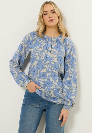 Blondynka w beżowej bluzce z długim rękawem w niebieski kwiatowy wzór, w połączeniu z niebieskimi dżinsami, stoi na tle gładkiej ściany.