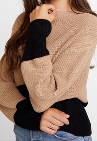 Gestrickter Pullover mit beigem Körper und schwarzen Ärmeln, ausgestattet mit Rippenstruktur und rundem Ausschnitt. Das Design umfasst kontrastierende Farbblöcke.