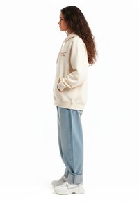 Felpa beige con cappuccio e tasca frontale, caratterizzata da testo rosso, abbinata a jeans baggy azzurri e sneakers bianche.