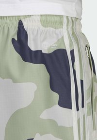 Pantaloni corti mimetici verdi e bianchi con accenti neri, vita elastica, texture a griglia e un logo che recita "FINE AI RIFIUTI DI PLASTICA" sul lato.