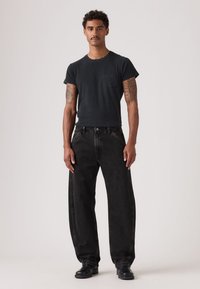 Μαύρο κοντομάνικο μπλουζάκι με τσέπη στο στήθος, συνδυασμένο με φαρδιά μαύρα τζιν τζιν. Το μοντέλο φοράει μαύρα μποτάκια· τατουάζ φαίνονται στους βραχίονες.