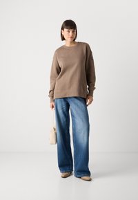Brun genser med rund hals og ribbestrikket kant, kombinert med vide blå denimbukser. Modellen holder en liten beige veske.
