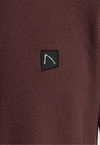 Tissu texturé bordeaux avec un motif gaufré subtil, présentant un petit patch noir avec un dessin de ligne blanche, positionné sur le côté gauche.