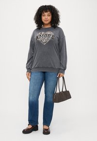 Sweatshirt cinzenta com um coração com estampa de leopardo e texto "AMORE", combinada com jeans azuis e sapatos castanhos, segurando uma pequena mala castanha.