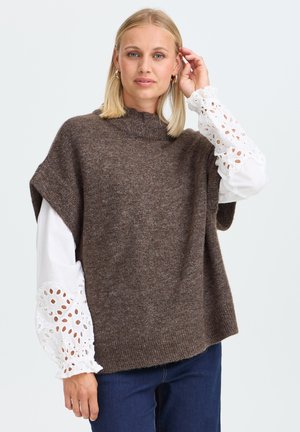 Femme aux cheveux blonds portant un pull sans manches en tricot marron par-dessus un chemisier blanc avec des manches en dentelle à œillets et un jean bleu, posant avec la main près du visage.