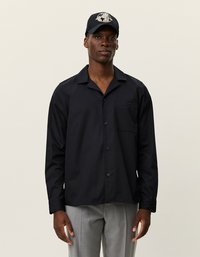 Camisa negra de botones con un bolsillo en el pecho, con un cuello relajado. Combinada con una gorra negra que muestra un logotipo gráfico y pantalones grises.