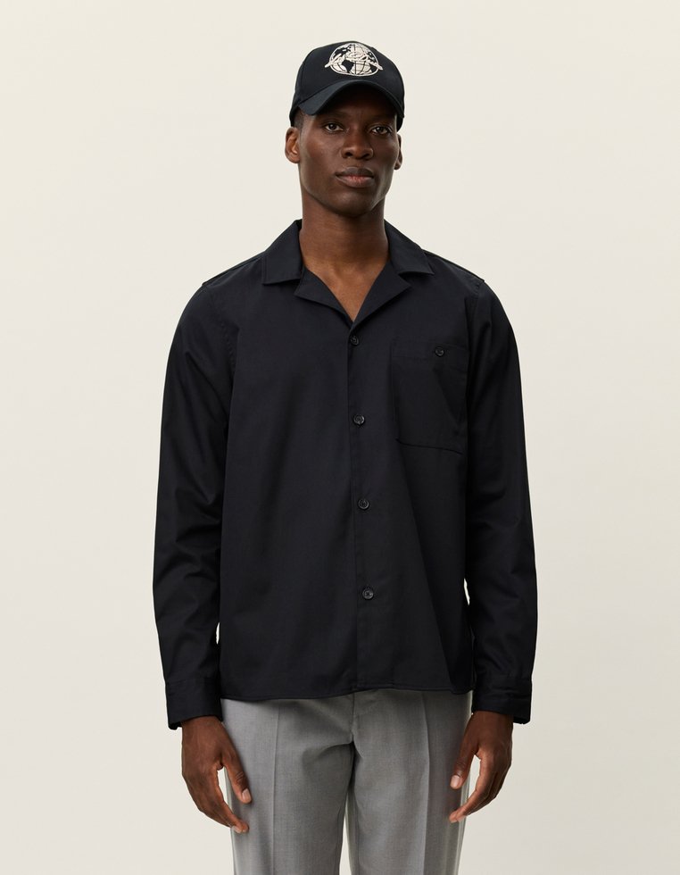 Camisa negra de botones con un bolsillo en el pecho, con un cuello relajado. Combinada con una gorra negra que muestra un logotipo gráfico y pantalones grises.